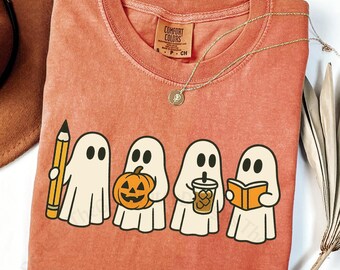 May include: A yam-colored Comfort Colors t-shirt featuring a graphic of four cartoon ghosts. One ghost holds a pencil, another a pumpkin, a third a drink, and the last a book. A gold necklace is draped over the shirt.
