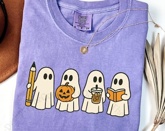 May include: A violet Comfort Colors t-shirt featuring a line of cartoon ghosts, each holding a different item: a pencil, a pumpkin, a coffee cup, and a book. The shirt has a crew neck and a tag that reads "Comfort Colors". A gold necklace and a brown hat are also visible.