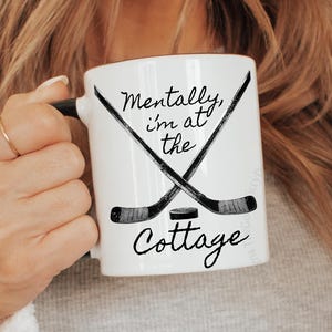 以下が含まれることがあります： 黒いハンドルが付いた白いセラミックマグ。テキスト「Mentally, I'm at the Cottage」と交差したホッケースティックとパックが表示されています。 マグカップは長い金髪の人が持っています。