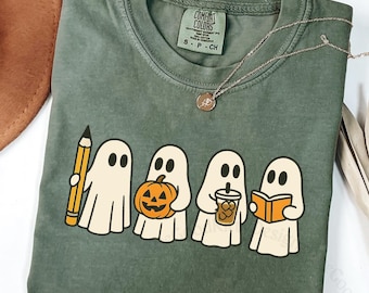 May include: Moss green t-shirt featuring four cartoon ghosts, each holding a different item: a pencil, a jack-o'-lantern, a coffee drink, and a book. The shirt has a crew neck and a Comfort Colors tag. A gold necklace is draped across the shirt.