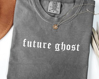 May include: A gray Comfort Colors t-shirt with the words "future ghost" in white gothic font. A gold necklace with a pendant rests on the shirt. A brown hat and dried plant are in the background.