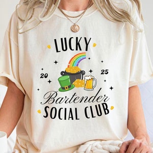 May include: A white t-shirt with the text "Lucky Bartender Social Club" in black. The design includes a rainbow, a pot of gold, a leprechaun hat, and a beer mug.