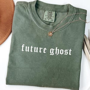May include: A moss green t-shirt with the words "future ghost" printed in white gothic font. A gold necklace with a pendant rests on the shirt. The shirt is folded and placed on a white surface, with a brown hat and dried plant in the background.