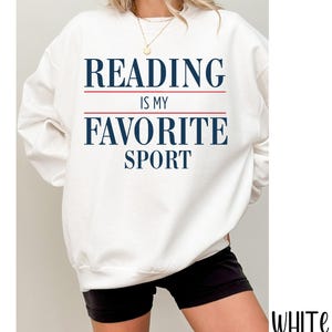 May include: White sweatshirt with the text "READING IS MY FAVORITE SPORT" in navy blue, with red lines separating the words. The sweatshirt is a casual style, perfect for book lovers.