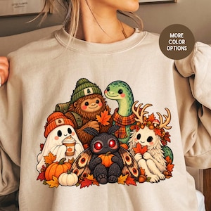 May include: A beige crewneck sweatshirt featuring a colorful graphic print of a group of cartoon creatures including a ghost, a yeti, a moth, and a dinosaur, all surrounded by autumn leaves, pumpkins, and a coffee cup.