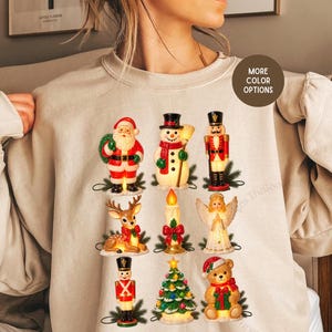 May include: A beige crewneck sweatshirt featuring a graphic print of nine vintage-style Christmas figures arranged in a grid. The figures include Santa Claus, a snowman, a nutcracker, a reindeer, a candle, an angel, a soldier, a tree, and a bear.