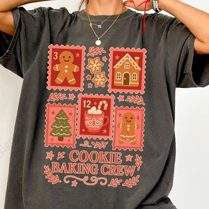 May include: Dark gray t-shirt featuring a festive design. The design includes gingerbread men, houses, and a Christmas tree, all within postage stamp-like frames. The text "COOKIE BAKING CREW" is displayed in a decorative font, perfect for the holiday season.
