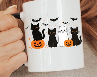 May include: White ceramic mug with a black handle featuring a Halloween design. The design includes black cats, orange pumpkins, and black bats. The mug is held by a person.
