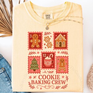 May include: A light yellow t-shirt with a graphic of gingerbread men, a gingerbread house, and a Christmas tree. The text "COOKIE BAKING CREW" is printed below the graphics. The shirt is paired with blue jeans and white shoes.