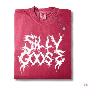 May include: A folded chili-coloured t-shirt with the words "SILLY GOOSE" in a white, stylised font. A silver-coloured necklace with a circular pendant rests on the shirt. The shirt has a Comfort Colors tag.