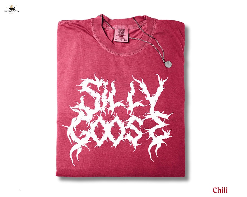 May include: A folded chili-coloured t-shirt with the words "SILLY GOOSE" in a white, stylised font. A silver-coloured necklace with a circular pendant rests on the shirt. The shirt has a Comfort Colors tag.