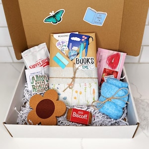 May include: A white gift box filled with a variety of items, including a blue washcloth, a brown flower-shaped coaster, a small package of Biscoff cookies, a small bag of Swiss Miss hot chocolate mix, a book-themed sticker, and a small bag of strawberry-flavored lip balm.