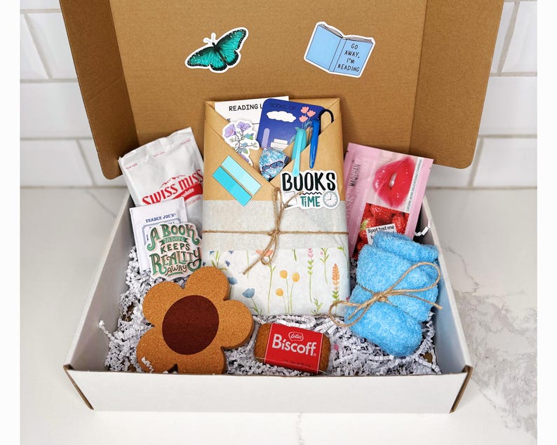 May include: A white gift box filled with a variety of items, including a blue washcloth, a brown flower-shaped coaster, a small package of Biscoff cookies, a small bag of Swiss Miss hot chocolate mix, a book-themed sticker, and a small bag of strawberry-flavored lip balm.