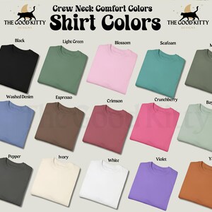 May include: A display of folded crew neck t-shirts in various colors, including black, light green, pink, and blue. The image includes the text "Crew Neck Comfort Colors" and "Shirt Colors", along with the brand logo "THE GOOD KITTY DESIGNS".