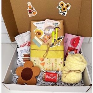 May include: A white gift box filled with a variety of items, including a book, a yellow towel, a brown flower-shaped coaster, a chocolate bar, and a small bag of candy. The box is decorated with a brown and white patterned paper and a yellow ribbon.