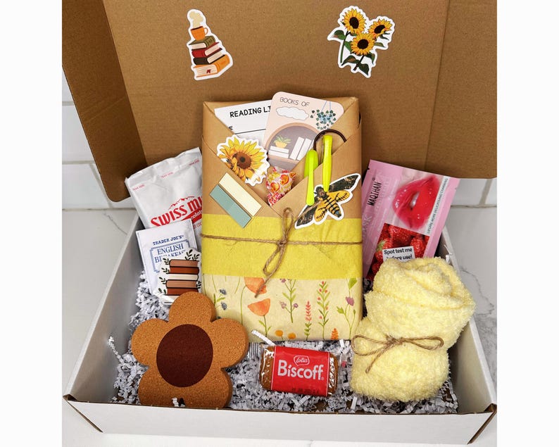 May include: A white gift box filled with a variety of items, including a book, a yellow towel, a brown flower-shaped coaster, a chocolate bar, and a small bag of candy. The box is decorated with a brown and white patterned paper and a yellow ribbon.