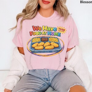 May include: A light pink t-shirt with the text "We Have Food At Home 1998 Edition" and a graphic of a TV dinner. The shirt is made of a soft material and is a casual wear item.