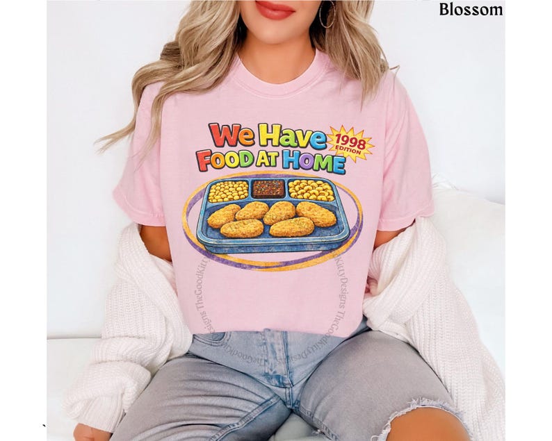 May include: A light pink t-shirt with the text "We Have Food At Home 1998 Edition" and a graphic of a TV dinner. The shirt is made of a soft material and is a casual wear item.