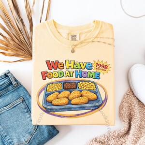 May include: A light yellow t-shirt with the text "We Have Food At Home 1998 Edition" and a graphic of a TV dinner. The shirt is laid flat with blue jeans, white sneakers, and a necklace.