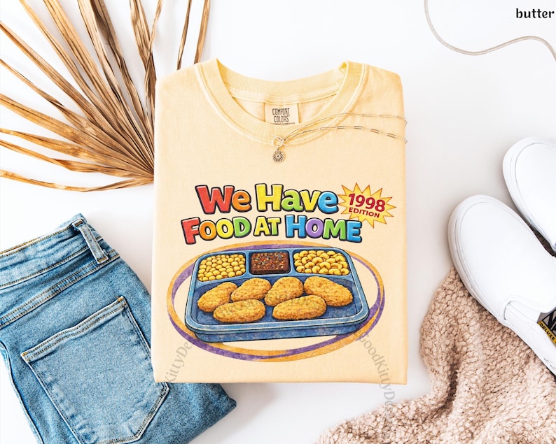 May include: A light yellow t-shirt with the text "We Have Food At Home 1998 Edition" and a graphic of a TV dinner. The shirt is laid flat with blue jeans, white sneakers, and a necklace.