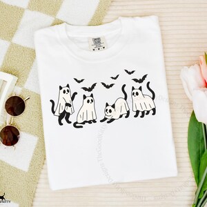 May include: White t-shirt featuring a Halloween-themed design. The graphic includes four black cats, some wearing ghost costumes, and several black bats flying above them. The word "white" is printed at the top right.