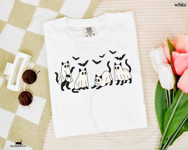 May include: White t-shirt featuring a Halloween-themed design. The graphic includes four black cats, some wearing ghost costumes, and several black bats flying above them. The word "white" is printed at the top right.