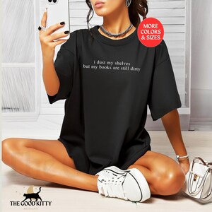 May include: Black t-shirt with the text "I dust my shelves but my books are still dirty" printed on the front. The shirt is being modeled by a person.