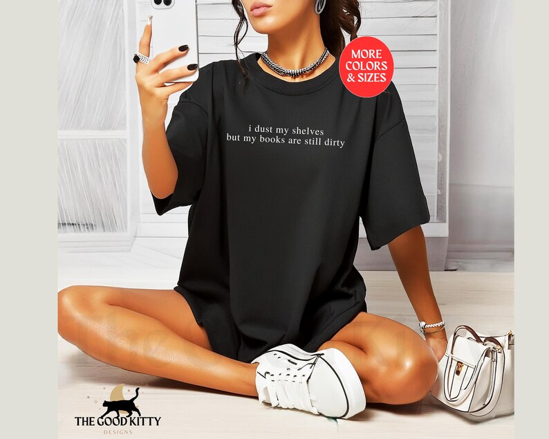 May include: Black t-shirt with the text "I dust my shelves but my books are still dirty" printed on the front. The shirt is being modeled by a person.