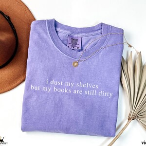 May include: A lavender colored t-shirt with the text "i dust my shelves but my books are still dirty" printed in white.