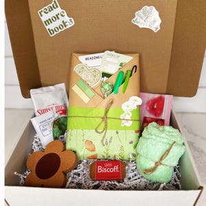 May include: A white box filled with a variety of items, including a green and brown book with a floral design, a green towel, a brown flower-shaped object, a small package of cookies, and a bag of Swiss chocolate. The box is decorated with a sticker that says "read more books".
