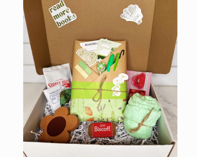May include: A white box filled with a variety of items, including a green and brown book with a floral design, a green towel, a brown flower-shaped object, a small package of cookies, and a bag of Swiss chocolate. The box is decorated with a sticker that says "read more books".