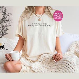 May include: A white t-shirt with the text "I dust my shelves but my books are still dirty" printed on it. The shirt is being worn by a person sitting on a bed with a white blanket.