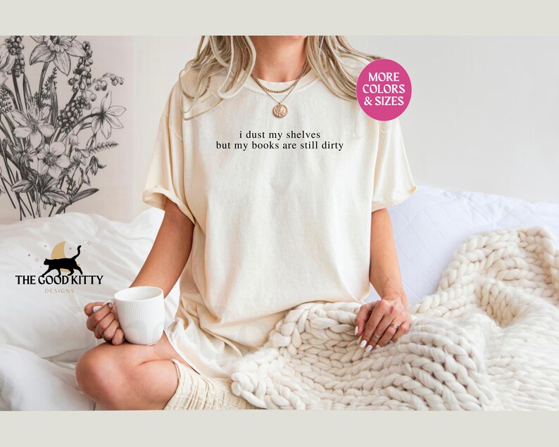May include: A white t-shirt with the text "I dust my shelves but my books are still dirty" printed on it. The shirt is being worn by a person sitting on a bed with a white blanket.