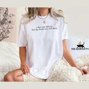 May include: White t-shirt with the text "I dust my shelves but my books are still dirty" printed on the front. The Good Kitty Designs logo is visible in the bottom right corner.