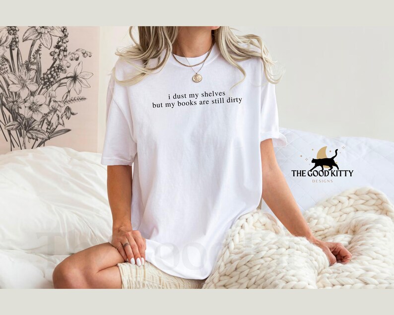 May include: White t-shirt with the text "I dust my shelves but my books are still dirty" printed on the front. The Good Kitty Designs logo is visible in the bottom right corner.