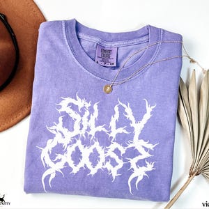 May include: A violet Comfort Colors t-shirt with the words "SILLY GOOSE" in a white, stylized font. A gold necklace with a pendant is draped across the shirt. A brown hat and dried palm fronds are in the background.