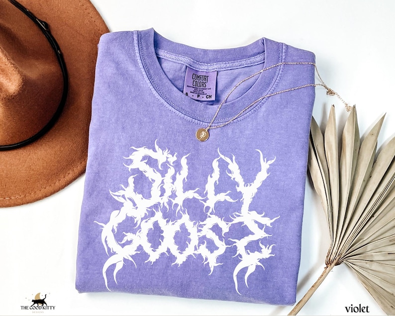 May include: A violet Comfort Colors t-shirt with the words "SILLY GOOSE" in a white, stylized font. A gold necklace with a pendant is draped across the shirt. A brown hat and dried palm fronds are in the background.