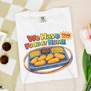 May include: White t-shirt with the text "We Have Food At Home 1998 Edition" in colorful letters. The shirt features a cartoon illustration of a TV dinner with chicken nuggets, peas, and other food items. The Good Kitty Design logo is visible.