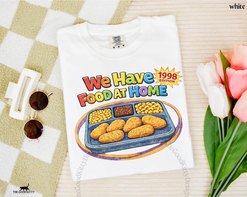 May include: White t-shirt with the text "We Have Food At Home 1998 Edition" in colorful letters. The shirt features a cartoon illustration of a TV dinner with chicken nuggets, peas, and other food items. The Good Kitty Design logo is visible.