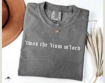 May include: A gray Comfort Colors t-shirt with the phrase "'twas the 'tism m'lord" printed in white. A gold-colored necklace with a pendant rests on the shirt. A brown hat and dried plant stems are in the background.