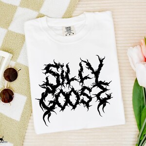 May include: A white t-shirt featuring the text "SILLY GOOSE" in a black, ornate font. The shirt is folded and placed on a beige and white checkered surface. Sunglasses and a hair clip are next to the shirt, with a bouquet of tulips.