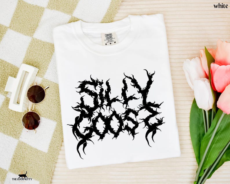 May include: A white t-shirt featuring the text "SILLY GOOSE" in a black, ornate font. The shirt is folded and placed on a beige and white checkered surface. Sunglasses and a hair clip are next to the shirt, with a bouquet of tulips.