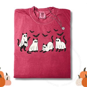 May include: A red t-shirt featuring a Halloween design with four cats dressed as ghosts and black bats. The shirt has a crew neck and short sleeves. The text "Chili" is visible in the upper right corner.