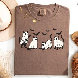 May include: Espresso-colored t-shirt featuring a playful Halloween design. The graphic includes four black cats dressed as ghosts, with bats flying above. A gold necklace and a hat are next to the shirt.
