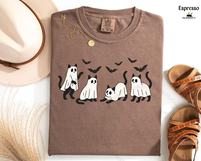 May include: Espresso-colored t-shirt featuring a playful Halloween design. The graphic includes four black cats dressed as ghosts, with bats flying above. A gold necklace and a hat are next to the shirt.