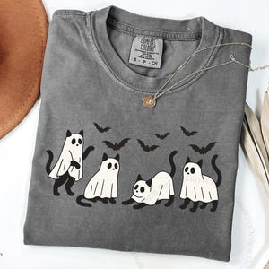 May include: A gray Comfort Colors t-shirt featuring a Halloween design. The shirt displays four cartoon cats dressed as ghosts, with black bat silhouettes above them. A silver necklace with a pendant is on the shirt.