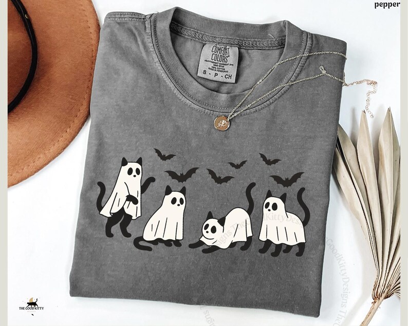 May include: A gray Comfort Colors t-shirt featuring a Halloween design. The shirt displays four cartoon cats dressed as ghosts, with black bat silhouettes above them. A silver necklace with a pendant is on the shirt.