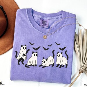 May include: A violet Comfort Colors t-shirt featuring a Halloween-themed design. The graphic includes four black cats, some wearing ghost costumes, and several black bats. A gold necklace with a pendant is draped on the shirt.