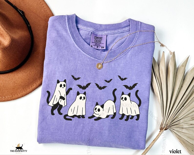 May include: A violet Comfort Colors t-shirt featuring a Halloween-themed design. The graphic includes four black cats, some wearing ghost costumes, and several black bats. A gold necklace with a pendant is draped on the shirt.