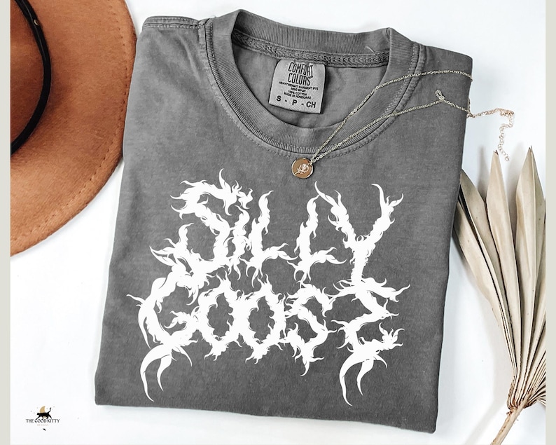May include: A grey Comfort Colors t-shirt with the words "SILLY GOOSE" in white, gothic lettering. A silver necklace with a small pendant rests on the shirt. A brown hat and dried leaves are in the background.
